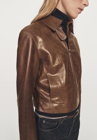 Brown leather cropped jacket with a glossy finish, featuring a collared neck, zip fastening, and black knit hem detail.
