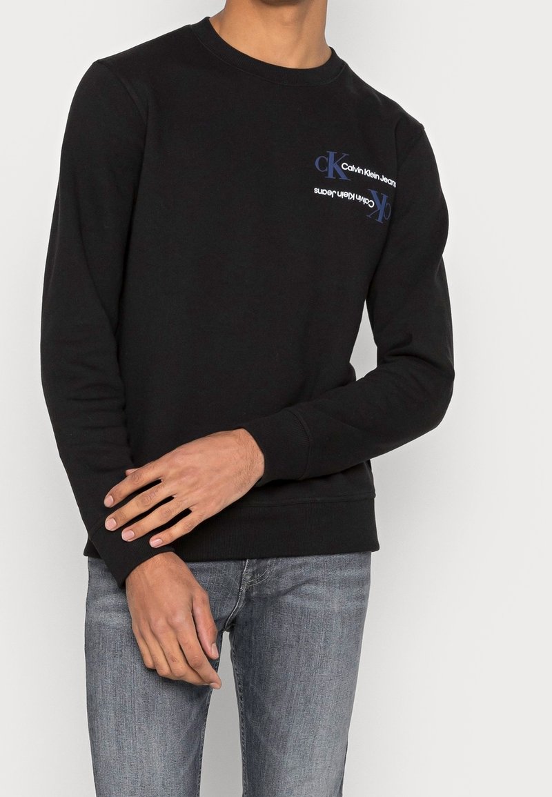 Černá mikina s kulatým výstřihem, žebrovanými manžetami a lemem. Na přední straně je modré logo "Calvin Klein Jeans". Měkký materiál z bavlněné směsi.
