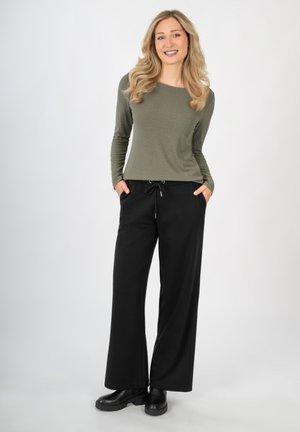 Langärmliges olivgrünes Oberteil mit einem strukturierten Muster, kombiniert mit schwarzen weiten Hosen mit elastischem Bund und seitlichen Taschen. Schwarze Stiefel.