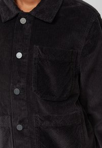 Schwarze Cordjacke mit Kragen, Fronttasche und Druckknöpfen. Der Stoff hat eine gerippte Textur, die die visuelle Tiefe verstärkt.
