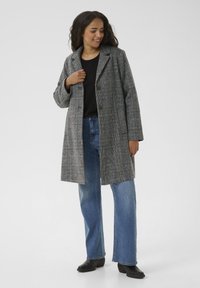 Grauer karierter Überrock mit Reverskragen, zwei Knöpfen und seitlichen Taschen, getragen über einem schwarzen Oberteil und blauen Jeans, kombiniert mit schwarzen Stiefeln.