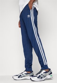 Plave sportske hlače od glatkog materijala, s tri bijele vertikalne pruge sa strane i Adidas logotipom sprijeda.