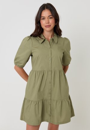 Mujer de cabello oscuro que lleva un vestido verde oliva con botones y mangas cortas, con una falda con volantes, de pie con las manos detrás de la espalda.