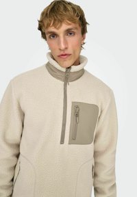 Villakangastakki vetoketjulla off-white-värissä, jossa on korkea kaulus, taskupatchi ja kontrastiväriset hiekanruskeat yksityiskohdat. Pinta on teksturoitu ja siinä on vetoketjuyksityiskohtia.