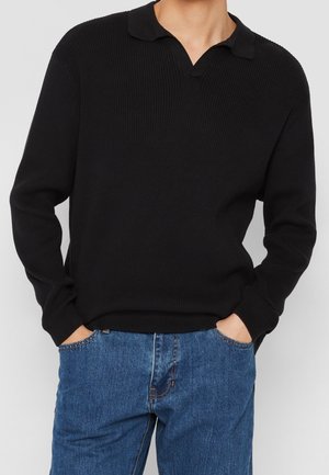 Sort langærmet ribstrikket sweater med slå-om-krave, båret med blå denimjeans, hænderne stukket i forlommerne, neutral baggrund.