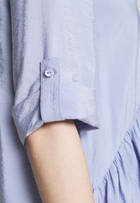 Světle fialová blůza se strukturovaným designem, s knoflíkovým manžetou a jemnou, zmačkanou texturou. Volánkový akcent po straně.