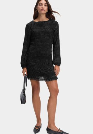 Femme portant une mini-robe noire texturée à manches longues, tenant un sac à main noir, et portant des ballerines pointues noires.