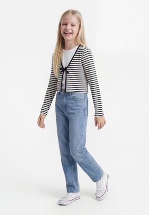 Chica sonriente con un cárdigan de rayas, camiseta blanca, vaqueros azules y zapatillas blancas, de pie frente a un fondo blanco liso.