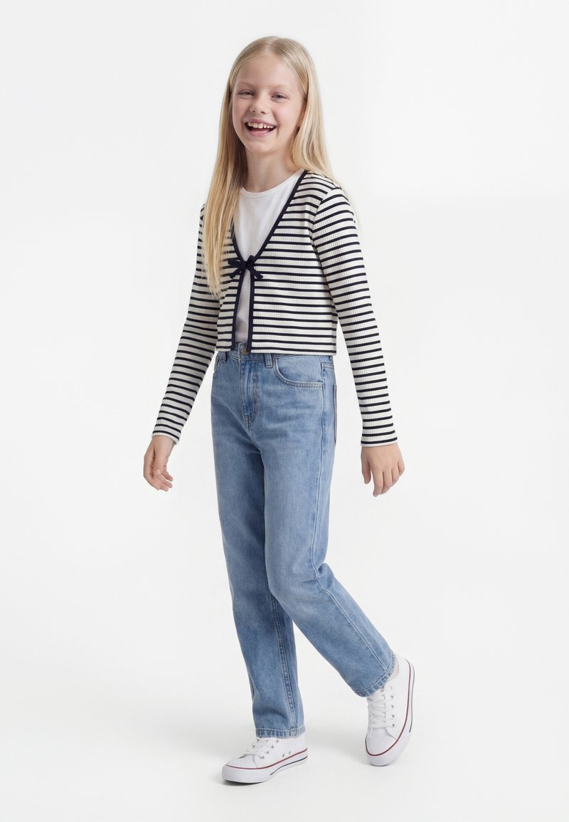 Uśmiechnięta dziewczyna ubrana w paskowany kardigan, białą koszulkę, niebieskie dżinsy i białe trampki, stojąca na jednolitym białym tle.