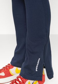 Ναυτικά παντελόνια φόρμας με λεία υφή, που διαθέτουν φερμουάρ στο πλάι και λεπτομέρεια λογότυπου. Συνδυασμένα με κόκκινες και κίτρινες αθλητικές τουαλέτες με πολύχρωμο σχέδιο.