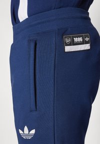 Mornarske hlače iz mehkega materiala, ki imajo logo značko z napisom "1886 Arsenal" in emblem Adidas s tremi listi v beli barvi. Vključene so stranske žepke.