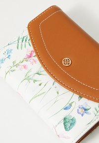 Portefeuille à motif floral en simili cuir blanc avec un rabat en simili cuir marron. Présente des accessoires en métal doré et des accents de couture blanche.
