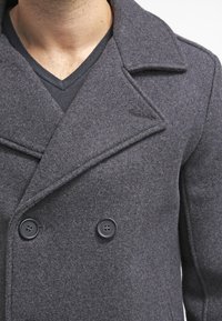 Palton gri din lână cu design dublu-două, cu rever mari fante și două nasturi negri. Materialul are o textură moale. Cămașă cu decolteu în V dedesubt.