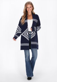 Cardigan blu navy con motivi geometrici bianchi, maniche lunghe, apertura frontale e vestibilità rilassata. Abbinato a jeans svasati in denim e stivali neri.