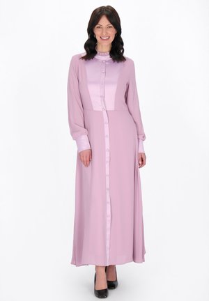 Mosolygó nő, hosszú pink gombos ruhában, fodros gallérral és mandzsettával, fehér háttér előtt állva.