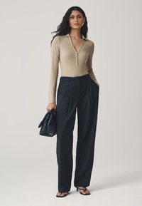 Camisa beige de manga larga con escote en V y detalles de botones, combinada con pantalones de talle alto en azul marino oscuro y un bolso negro tejido.