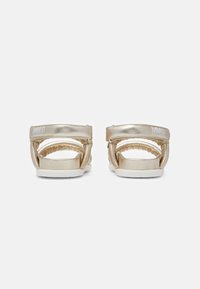 Ett par metalliska guldskor med vadderade remmar och vita sulor, sedda bakifrån mot en vit bakgrund.