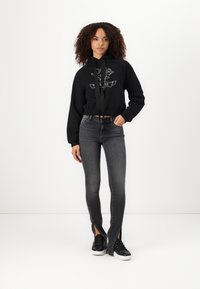 Black cropped hoodie with logo, paired with dark grey skinny jeans featuring side slits. Model wears black sneakers. Simple design and casual look.