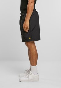 Svart shorts med elastisk midje og et smilende ansikt-logo som aksent. Brukt med hvite joggesko og ankelsokker. Flatt, lett stoff.