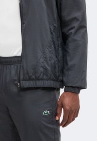 Gros plan d'une personne portant une veste noire zippée et un pantalon assorti avec le logo crocodile vert Lacoste sur la cuisse gauche.