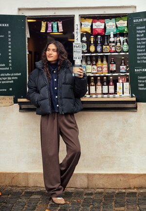 Femme en veste sombre et pantalon marron tenant une tasse de café, debout près d'un kiosque extérieur vendant des boissons et des encas avec des panneaux de menu visibles.