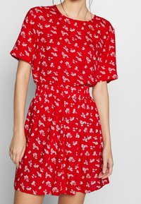 Donna che indossa un vestito rosso a maniche corte con un motivo floreale bianco e una cintura elastica, in piedi contro uno sfondo neutro.