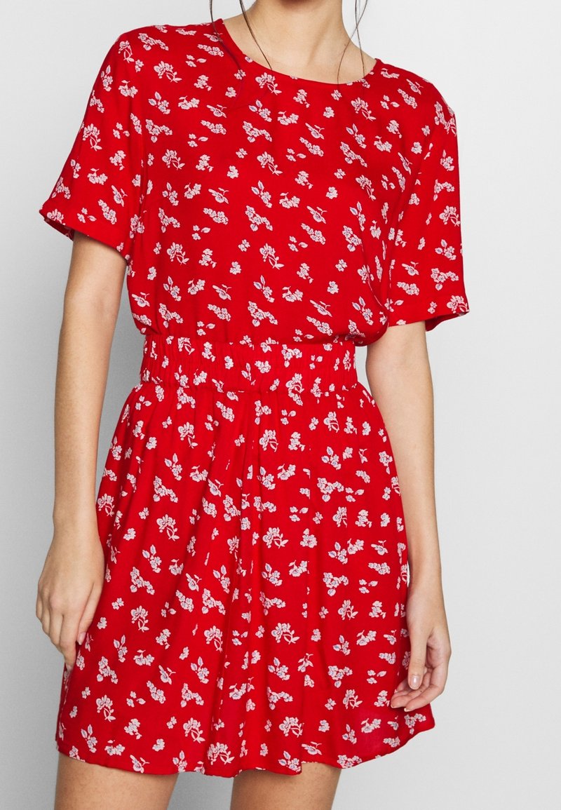 Donna che indossa un vestito rosso a maniche corte con un motivo floreale bianco e una cintura elastica, in piedi contro uno sfondo neutro.