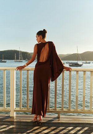 Femme en robe marron dos nu et talons, debout sur une terrasse en bois, surplombant une eau calme avec des voiliers et des collines lointaines.