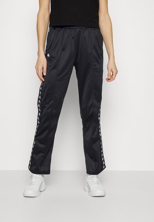 Person iført sorte træningsbukser med hvide logostriber ned langs siderne og hvide chunky sneakers, stående mod en ensfarvet hvid baggrund.