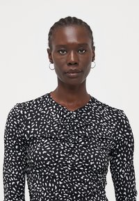 Haut noir à manches longues en tissu texturé avec un motif abstrait blanc, détail froncé à l'avant et encolure ronde.
