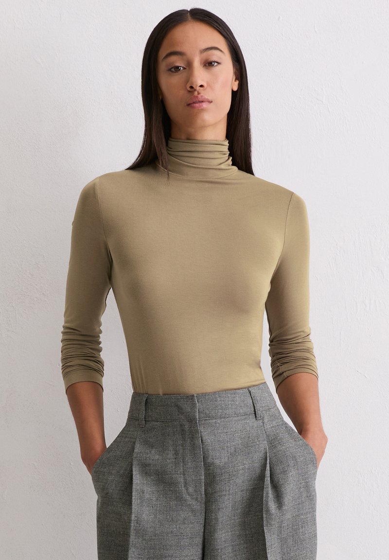 Frau mit langen, dunklen Haaren, die einen beigen Rollkragenpullover und graue hochgeschnittene, plissierte Hosen trägt, mit den Händen in den Taschen, stehend vor einer weißen Wand.