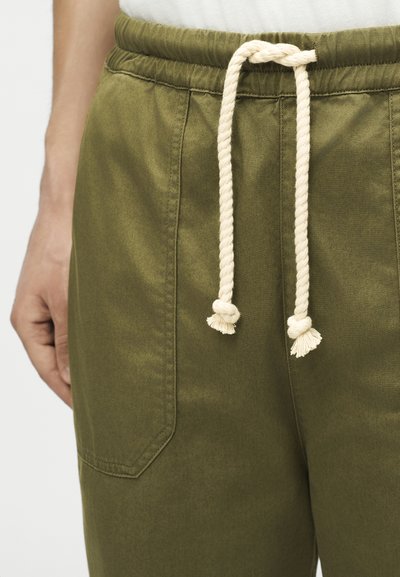 Κοντινό πλάνο σε χακί παντελόνι με μπροστινή τσέπη και εκρέμ κορδόνι στη μέση, φορεμένο από άτομο με λευκό τοπ.