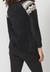 Schwarzer flauschiger Pullover mit strukturierten Schulterakzenten in Grau, Schwarz und goldfarbenem Zickzackmuster. Lange Ärmel und gerippter Saum.