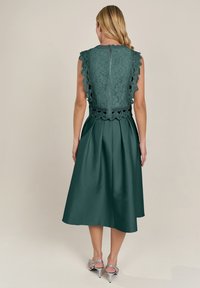 Grünes Spitzenkleid mit gewellten Ärmeln und Reißverschluss auf dem Rücken, ausgestattet mit einem vollen, strukturierten Rock und Taschen. Silberne Absätze vervollständigen den Look.