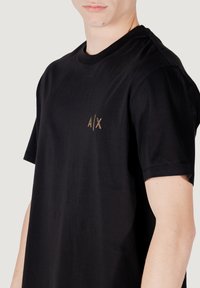 Zwarte katoenen T-shirt met een ronde hals en korte mouwen, voorzien van een klein gouden logo "A/X" op de linkerborst. Gladde textuur.