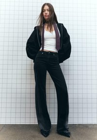 Crna unisex dukserica, bijela majica bez rukava, crne široke traperice visokog struka s bijelim šavovima, i crne cipele s upućenim vrhom. Detalj s vezicom u prugama.