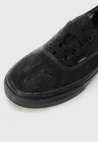 Fekete sneaker, csipkével díszített, kerek orrú, textúrált anyagból készült, fekete fűzőkkel és gumitalppal. A VANS logó az oldalon.