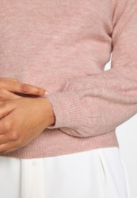 Jasnoróżowy sweter dziergany na drutach z prążkowanymi mankietami i dolnym obrębem, miękką teksturą, o luźnym kroju, noszony na białej bluzce.