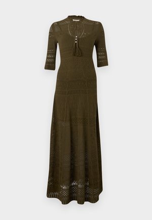 Žena u uzorkovanoj maslinasto zelenoj dugoj haljini, smeđim čizmama, sunčanim naočalama i velikom smeđom torbom na ramenu na jednobojnoj pozadini.