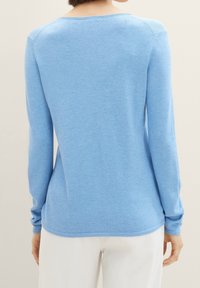 Frau trägt einen hellblauen langärmeligen Strickpullover und weiße Hosen, von hinten vor einem schlichten Hintergrund gezeigt.