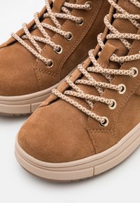 Brown suede high-top sneakers with textured surface, round toe, gold eyelets, and patterned tan/white laces on a light-colored sole.