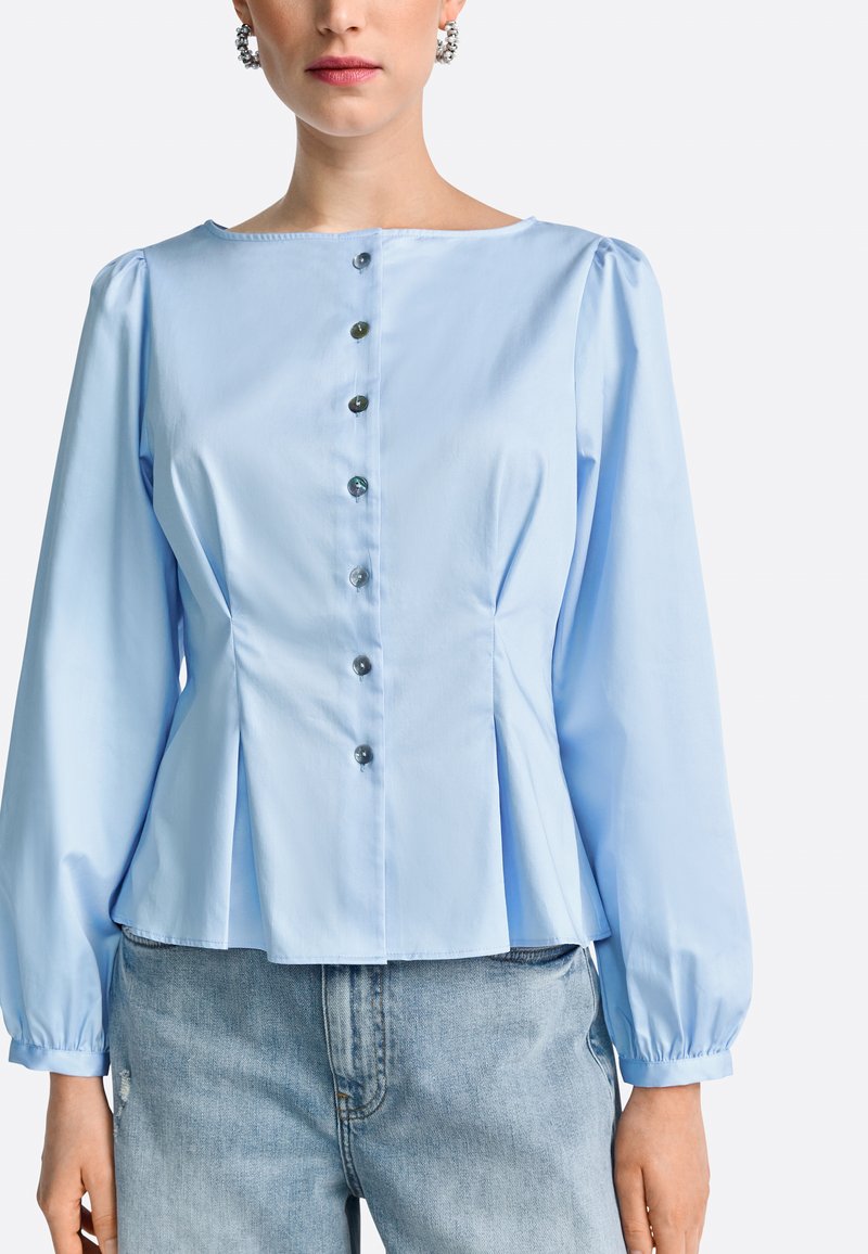 Light blue peplum blouse with long sleeves, round neckline, and button front detail. Smooth fabric with subtle sheen and gathered sleeves.