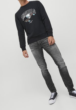 Persona que lleva una sudadera negra con un gráfico de calavera y monopatín, vaqueros grises desgastados y rotos, y zapatillas negras y blancas, inclinado hacia un lado.