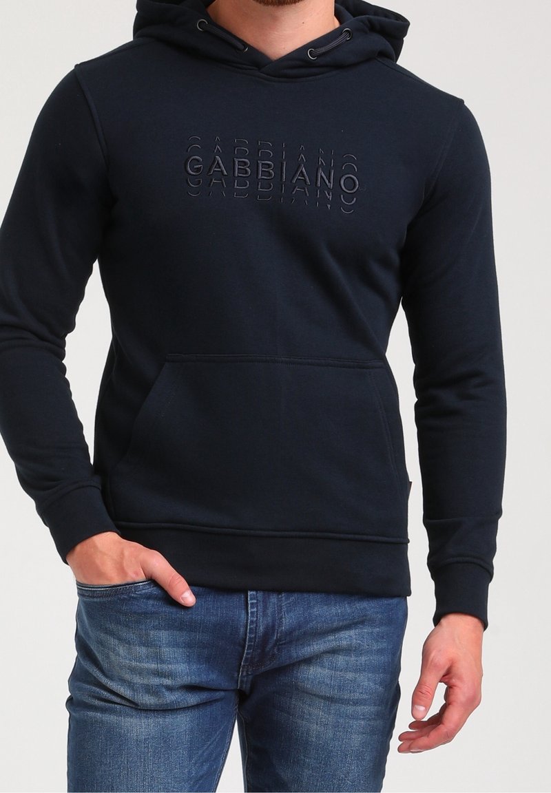 Homme portant un sweat à capuche bleu foncé avec le texte « "GABBIANO" », jean bleu, main gauche dans la poche, bras droit détendu, montré des épaules aux hanches.
