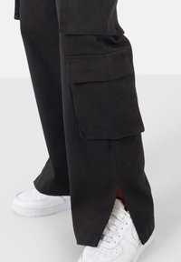 Pantalon cargo noir doté de deux grandes poches latérales, une coupe droite et des fentes latérales à l'ourlet, associé à des baskets blanches.