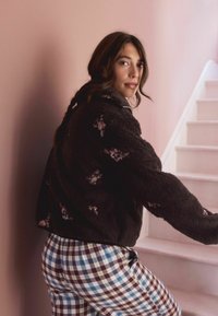 Rudas vilnos striukė su gėlių siuvinėjimu, derinama su mėlynomis ir maroon juostuotomis kelnėmis. Fone matyti švelni rožinė siena ir baltos laiptų pakopos.