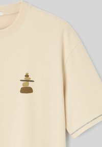 T-shirt beige à manches courtes avec un petit graphique de pierres empilées en nuances de marron et vert sur le côté gauche de la poitrine.