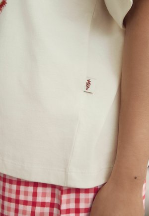 Nahaufnahme einer Person, die ein weißes Hemd mit einem kleinen roten Blatt-Logo und rot-weiß karierten Hosen trägt.