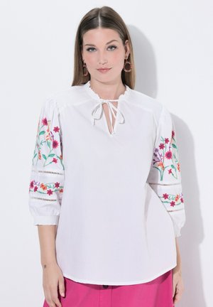 Femme portant un chemisier blanc avec broderie florale sur des manches trois-quarts bouffantes et un détail à nouer au col, associé à un bas rose vif.