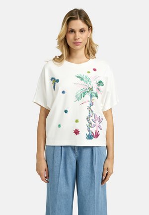 Frau trägt ein weißes, locker sitzendes T-Shirt mit bunter tropischer Blumen- und Vogelstickerei, kombiniert mit hochtaillierten hellblauen Jeans mit weitem Bein.